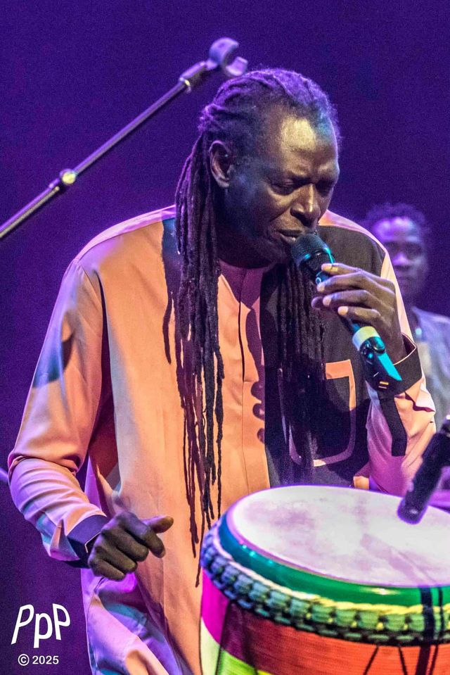 Zale Seck chante au micro derrière son djembé, sous une lumière violette.