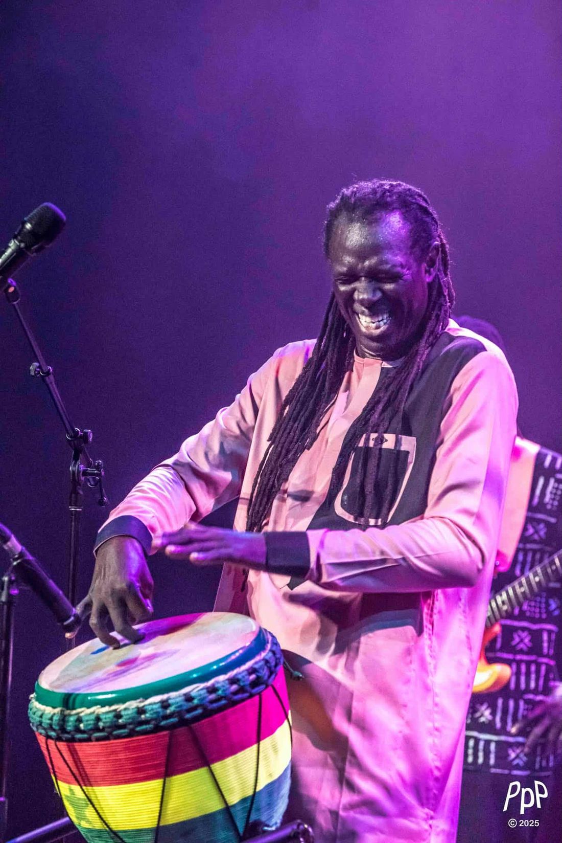 Zale Seck joue du djembé en riant, en boubou rose, sur scène.