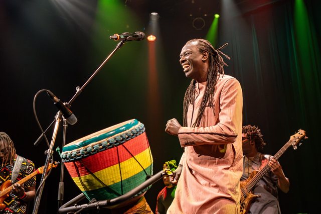Zale Seck rit sur scène, poing fermé, devant un dundun aux couleurs panafricaines.